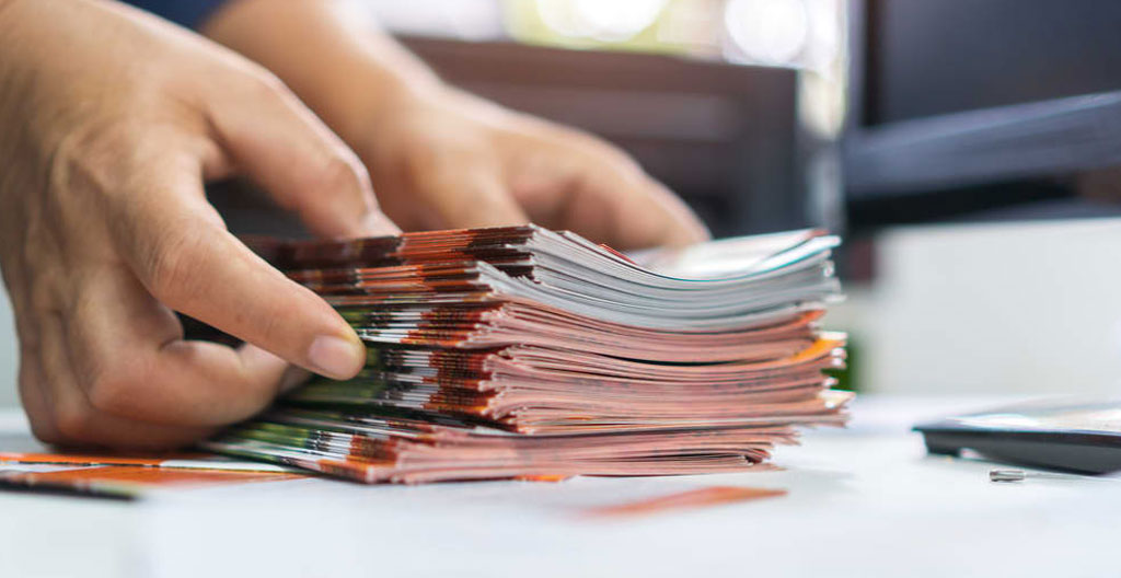 Extreme close-up of hands holding a printed brand guidelines document next to a laptop displaying a video storyboard, shallow depth of field